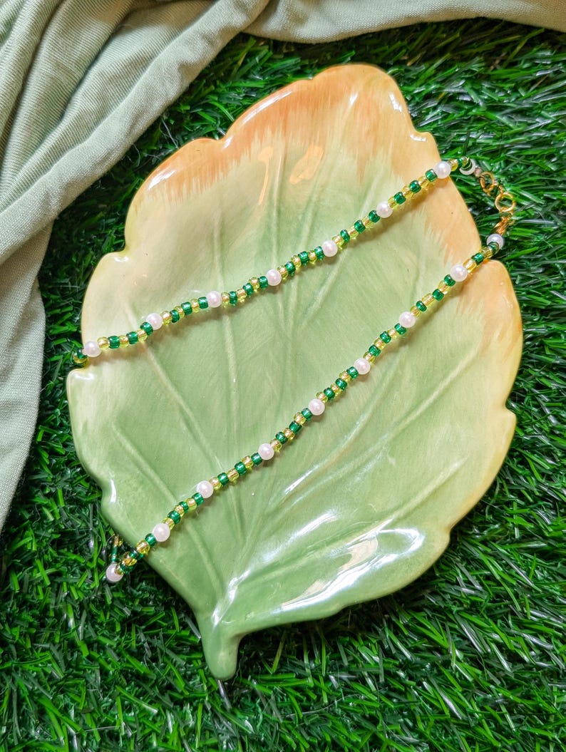 May include: A delicate gold chain necklace with alternating green and white beads. The necklace is displayed on a green leaf-shaped plate.