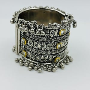 May include: Silver cuff bracelet with detailed floral and geometric patterns. The bracelet has small dangling silver beads and square gold-coloured accents. A detailed piece of jewellery.