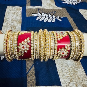 May include: A collection of red and gold bangles, adorned with intricate floral and geometric patterns. The bangles feature embedded gemstones and a polished finish, creating a luxurious and ornate appearance. The bangles are displayed on a white cylindrical surface.