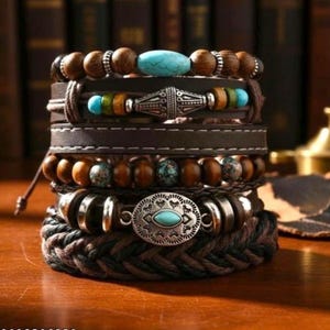 May include: A multi-layered bracelet stack featuring brown wooden beads, turquoise-colored stones, dark brown leather straps with white stitching, silver-toned metal spacers, and a central oval silver-toned charm with a turquoise inlay.