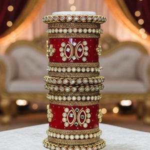 May include: A stack of ornate, red and gold bangles. The bangles are adorned with intricate gold detailing and sparkling gemstones. The design includes floral and geometric patterns, creating a luxurious and festive appearance. The bangles are stacked on a white surface.