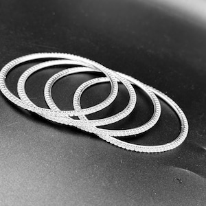 May include: Four silver-colored, circular bangles with a continuous line of small, sparkling stones. The bangles are arranged in a slightly overlapping pattern against a dark background, creating a display of jewelry.