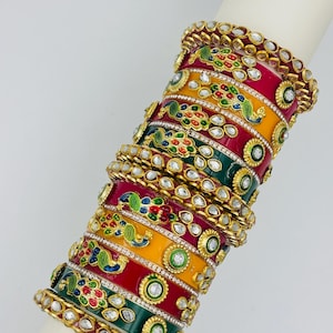 Op de afbeelding: Een stapel kleurrijke armbanden met ingewikkelde ontwerpen. De armbanden hebben rode, gele en groene banden, versierd met pauwmotieven en sprankelende edelstenen. Goudkleurige metalen accenten geven de sieraden een vleugje elegantie.