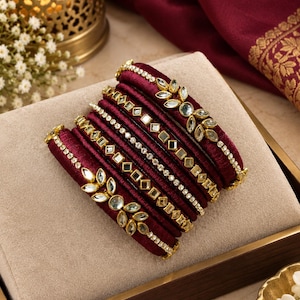 May include: A set of six deep maroon silk-wrapped bangles featuring gold-toned metal accents. The bangles are decorated with clear, faceted glass stones in leaf-shaped and square settings, along with thin rows of small, sparkling clear crystals.