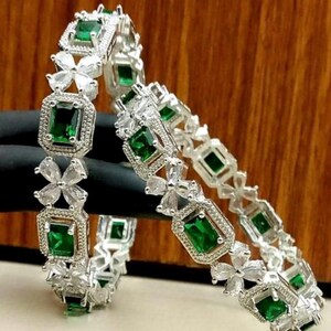 May include: A pair of silver-toned bangles featuring alternating emerald green rectangular stones and clear white floral-shaped accents. The metal settings are detailed with small, textured beads surrounding each of the vibrant green stones.