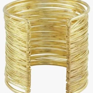 May include: A wide cuff bracelet constructed from multiple strands of thin, gold-toned wire wrapped horizontally around a circular frame. The design features an open gap at the front, showcasing the layered, textured appearance of the metallic wire.