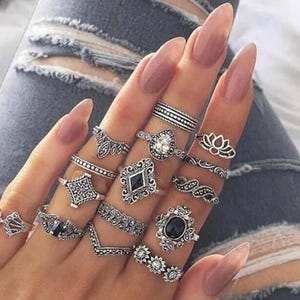 Puede incluir: Una colección de quince anillos de tono plateado usados en los dedos, con varios diseños que incluyen patrones de filigrana, formas geométricas, una piedra central clara y piedras ovaladas negras con detalles metálicos oxidados.