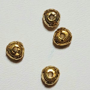 May include: Four gold-colored beads with a textured, floral-like design. Each bead has a central hole. The beads are scattered on a white surface, showcasing their intricate details and metallic sheen. These beads are suitable for jewelry making.