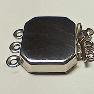 May include: A silver-toned, multi-strand jewelry clasp. The clasp has a rectangular, beveled design with three loops on each side for attaching strands. The clasp is made of a shiny, reflective metal.