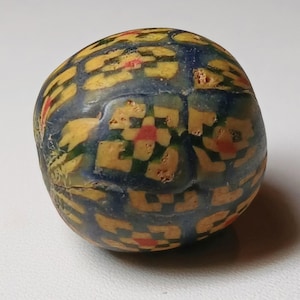 May include: A rounded, patterned glass bead with a mosaic design. The bead features a base of deep blue with yellow and red floral and checkered patterns. The bead has a slightly irregular shape and a smooth, polished surface.