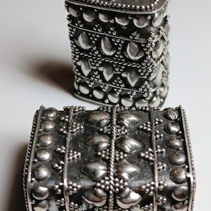 May include: Two ornate silver boxes. The taller box is rectangular with a hinged lid, and the shorter box is also rectangular. Both are decorated with raised floral and dot patterns, and are likely used for jewelry or small items.