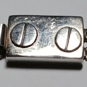 May include: A silver-toned jewelry clasp with a rectangular body and two circular screw heads. The clasp has a hook on one end and two loops on the other for attachment. The metal has a slightly worn appearance.