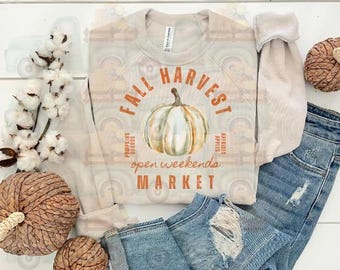 Può includere: Una felpa beige con la scritta "FALL HARVEST MARKET" e una grafica di zucca ad acquerello. Le maniche sono arrotolate e la felpa è abbinata a jeans e zucche decorative. Il testo include anche "Pumpkins, Hayrides, Apples, Open Weekends".