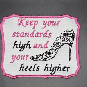 May include: Embroidered patch with a white background and a pink border. The patch features the text "Keep your standards high and your heels higher" in pink and black. A black high heel shoe with floral details is also present.