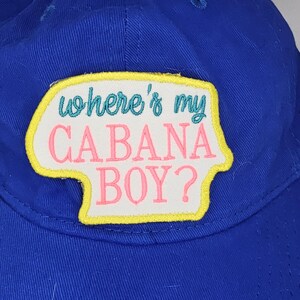 May include: A blue baseball cap with a patch that reads "where's my CABANA BOY?" in teal and pink lettering. The patch has a white background with a yellow border. The cap is made of a durable fabric.