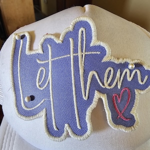 May include: White trucker hat with a large, embroidered patch. The patch is a light purple color with the words "let them" in white script, and a small pink heart. The hat has a mesh back.