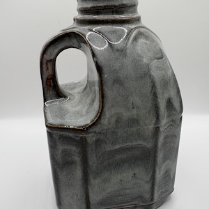 Puede incluir: Una jarra de cerámica hecha a mano con un esmalte gris. La jarra tiene un cuerpo rectangular, un asa redondeada y un cuello estrecho. El esmalte presenta variaciones de color, lo que añade encanto rústico. Ideal para la decoración del hogar.