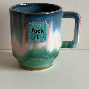 Könnte beinhalten: Eine Keramik-Tasse mit blauer, rosa und grüner Glasur. Die Tasse hat ein quadratisches Feld mit den Worten "fuck it" in Schwarz. Der Griff ist quadratisch und die Tasse steht auf einer weißen Oberfläche.
