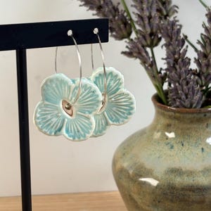 May include: Turquoise ceramic flower earrings with silver hoops. The earrings are displayed on a black stand. A vase of lavender is in the background. The earrings have a detailed floral design.