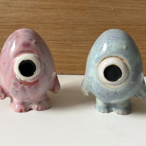 Może przedstawiać: Dwie ceramiczne figurki, jedna różowa i jedna niebieska, każda z jednym dużym okiem i małymi ramionami i stopami. Oko jest czarne z białym obramowaniem. Figurki mają fantazyjny, kreskówkowy design.