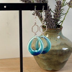 Puede incluir: Pendientes con detalles hexagonales plateados y colgantes azul-turquesa en forma de lágrima. Los pendientes se exhiben en un soporte negro, con un jarrón de lavanda en el fondo.