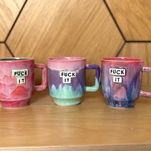 Pode incluir: Três canecas de cerâmica com acabamento brilhante, cada uma com uma placa branca quadrada com o texto preto "FUCK IT" centralizado na frente. As canecas exibem gradientes de rosa, roxo e turquesa, e cada uma tem uma alça quadrada.