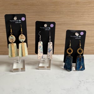 May include: Three pairs of handmade earrings on display. The earrings feature various designs, including geometric shapes and circular accents. Each pair is attached to a black card with the text "Handmade with love". The earrings are displayed on clear stands.