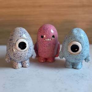 Könnte beinhalten: Drei Keramik-Monsterfiguren. Zwei sind hellblau mit dunkelblauen Sprenkeln und einem großen Auge. Die mittlere Figur ist rosa mit dunkelrosa Sprenkeln und einem kleinen Mund. Alle haben kleine Arme und Füße.