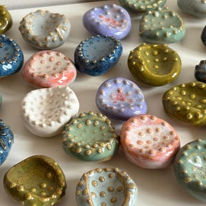 May include: A selection of small, handmade ceramic dishes in various colours, including blue, green, purple, and white. Each dish has a textured surface with small, raised dots around the rim and inside the bowl. The dishes are arranged on a white surface.
