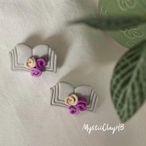 May include: Two grey clay book charms featuring purple and cream coloured flowers. The charms are handmade and have a delicate, whimsical design.