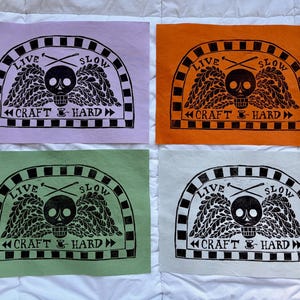 May include: Four rectangular prints in light purple, orange, green, and white, each featuring a black ink block print of a skull with wings, crossed knitting needles, and the text "LIVE SLOW" and "CRAFT HARD" inside a checkered arch border.
