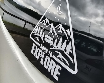 Könnte beinhalten: Weißer dreieckiger Aufkleber mit Bergmotiv, Mond und dem Schriftzug "BORN TO EXPLORE". Der Aufkleber befindet sich auf einer Fahrzeugscheibe, mit einem See und Himmel im Hintergrund.