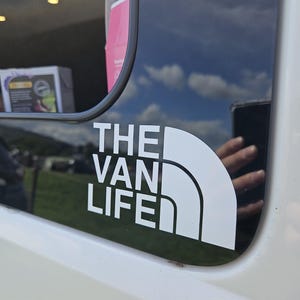 May include: White vinyl decal on a vehicle window. The decal reads "THE VAN LIFE" in a bold, sans-serif font, with a curved design element. The background shows a reflection of a blue sky with clouds and a hand.