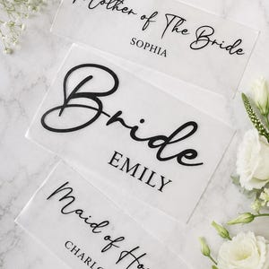 Könnte beinhalten: Drei rechteckige Schilder aus klarem Acryl mit schwarzer Beschriftung. Das oberste Schild zeigt Mother of the Bride SOPHIA, das mittlere Bride EMILY und das untere Maid of Honour CHARLOTTE in einer Kombination aus Schreib- und serifenloser Schrift.