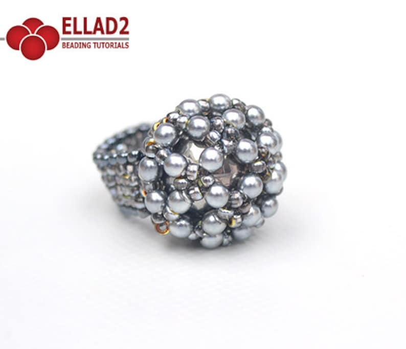 May include: A silver beaded ring with a round, textured design. The ring is made with small, round beads and has a slightly raised center.