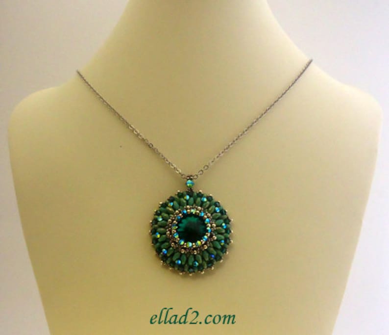 Op de afbeelding: Een zilveren ketting met een groene kralenhanger met een grote smaragdgroene edelsteen in het midden. De hanger is rond en heeft een symmetrisch ontwerp.