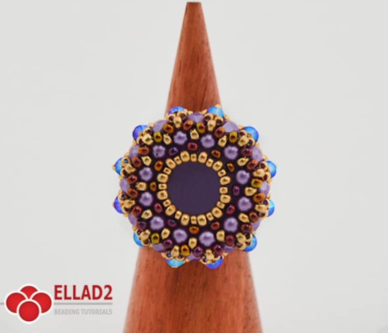 K&ouml;nnte beinhalten: Ein lilafarbener Perlenring mit einer gold- und braunen Mitte und blauen Akzenten. Der Ring wird auf einem h&ouml;lzernen Ringst&auml;nder pr&auml;sentiert.
