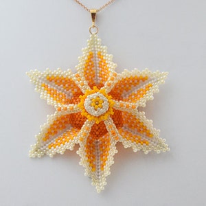 Puede incluir: Un collar con un colgante de flor de cuentas, con un diseño de estrella amarilla y blanca. El colgante está hecho de pequeñas cuentas redondas y cuelga de una cadena dorada.