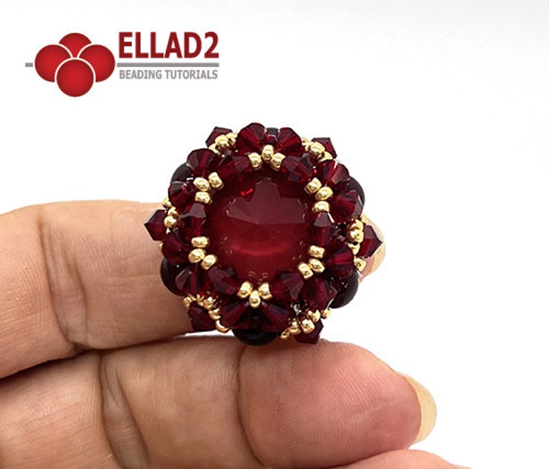 Może przedstawiać: Pierścionek z czerwonym kamieniem szlachetnym, z ozdobnym złotym obramowaniem i skupiskiem czerwonych kryształ&oacute;w wok&oacute;ł kamienia. Pierścionek jest na palcu.