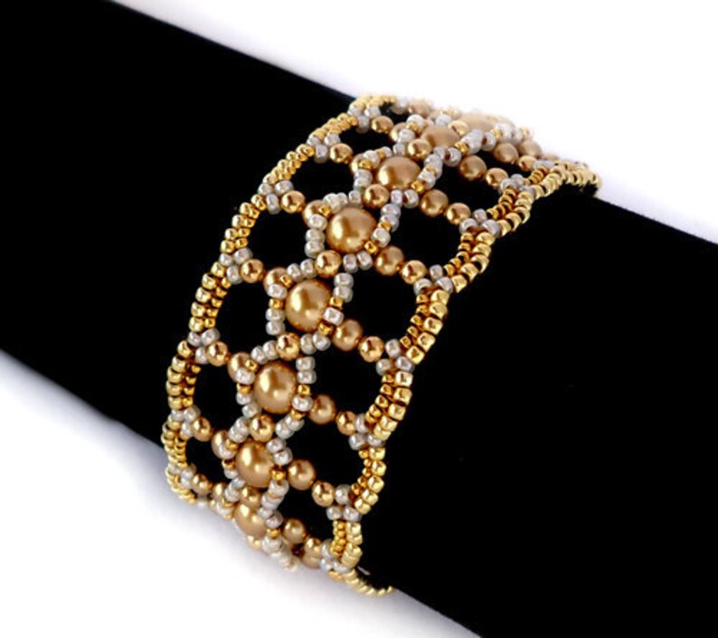 K&ouml;nnte beinhalten: Ein goldfarbenes und wei&szlig;es Armband mit geometrischem Muster. Das Armband ist aus kleinen Perlen und gr&ouml;&szlig;eren runden Perlen gefertigt.