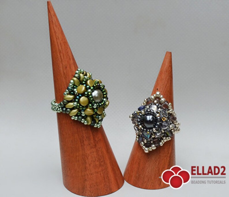 May include: Two beaded rings on wooden display stands. The ring on the left is green and gold with a pearl-like center bead. The ring on the right is silver and black with a dark gray center bead.