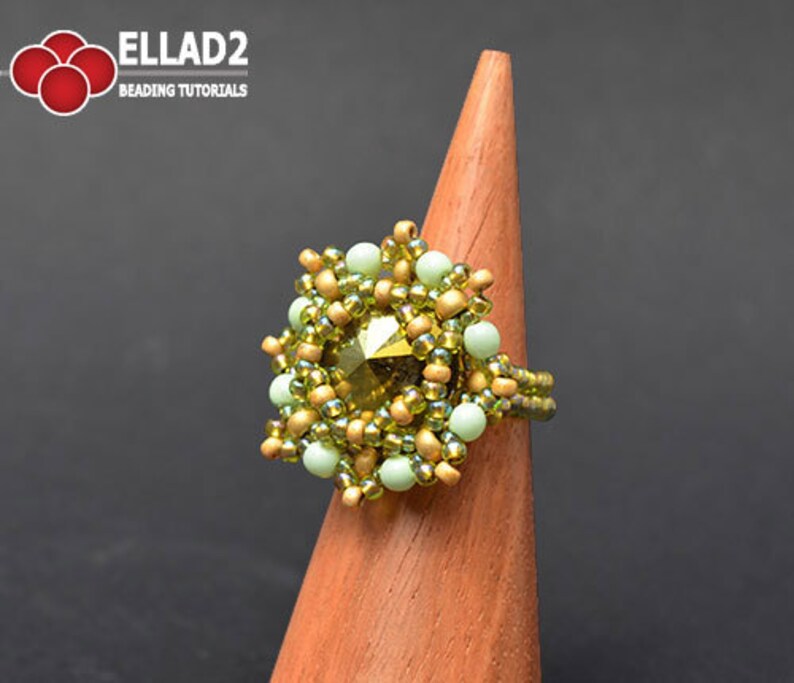 K&ouml;nnte beinhalten: Ein gr&uuml;ner und goldener Perlenring mit einem gro&szlig;en facettierten Edelstein in der Mitte. Der Ring wird auf einem h&ouml;lzernen Ringst&auml;nder pr&auml;sentiert.