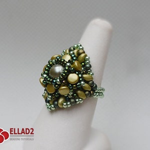May include: A green and gold beaded ring with a large pearl-like bead in the center. The ring is on a white ring mandrel.