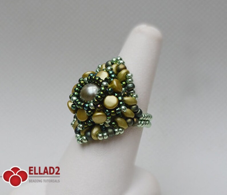 May include: A green and gold beaded ring with a large pearl-like bead in the center. The ring is on a white ring mandrel.