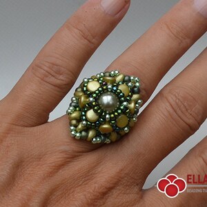 May include: A green and gold beaded ring with a pearl in the center. The ring is on a person's finger.