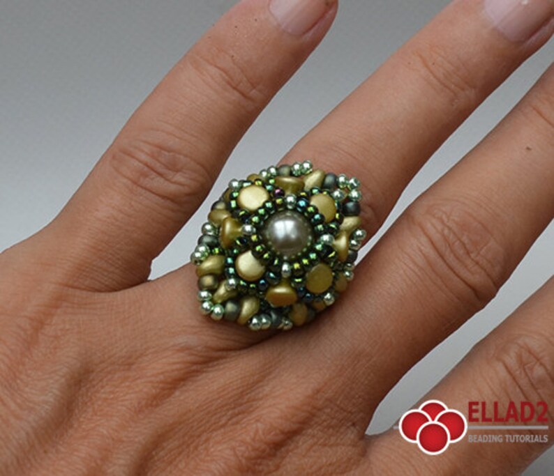 May include: A green and gold beaded ring with a pearl in the center. The ring is on a person's finger.