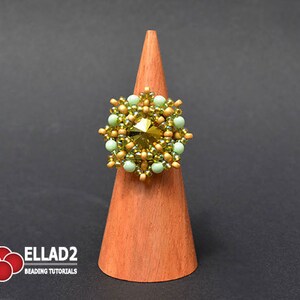 K&ouml;nnte beinhalten: Ein goldfarbener und gr&uuml;ner Perlenring mit einem gro&szlig;en, facettierten, klaren Kristall in der Mitte. Der Ring wird auf einem h&ouml;lzernen Ringst&auml;nder pr&auml;sentiert.