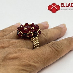 Może przedstawiać: Złoty pierścionek z czerwonym wzorem kwiatu z koralik&oacute;w. Pierścionek jest na palcu osoby.