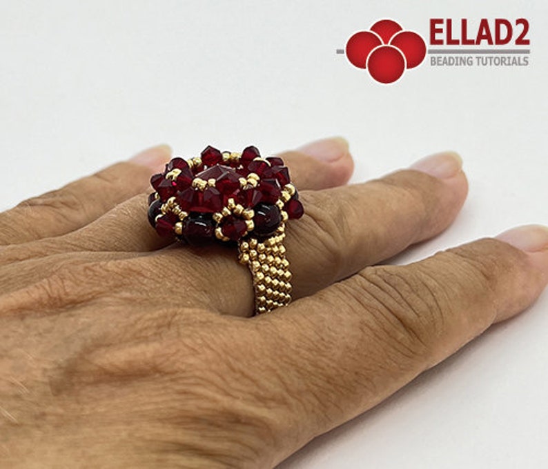 Może przedstawiać: Złoty pierścionek z czerwonym wzorem kwiatu z koralik&oacute;w. Pierścionek jest na palcu osoby.