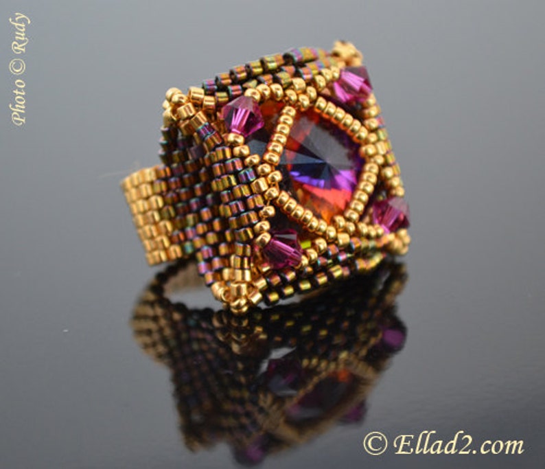May include: A square-shaped ring with a large, faceted, iridescent purple and orange gemstone surrounded by smaller, purple gemstones. The ring is made of gold-colored beads and has a woven design.